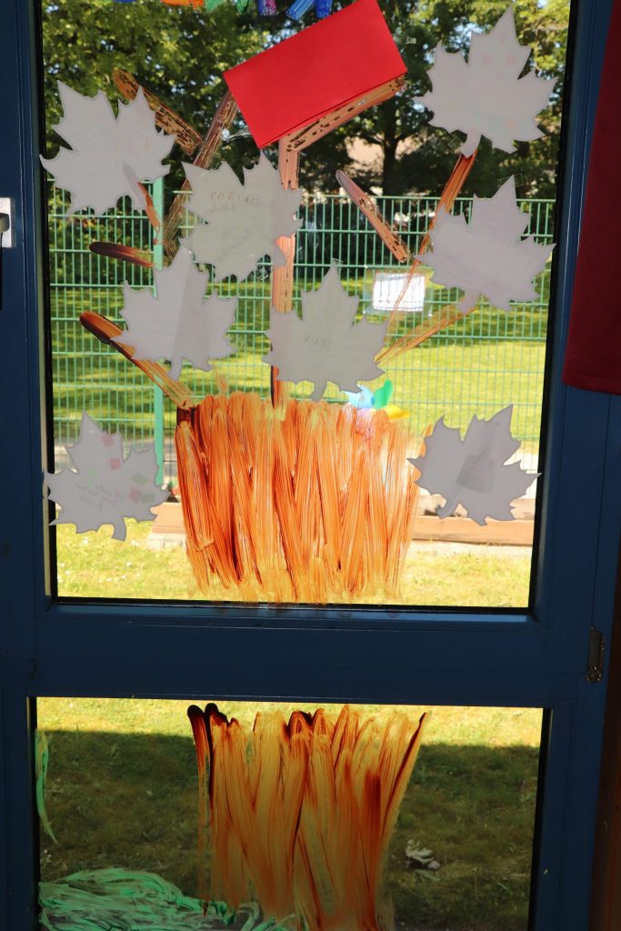 Ein Baum ist auf ein Fenster gemalt. An den Zweigen hängen Blätter aus Papier. Darauf stehen die Wünsche der Kinder und Eltern.